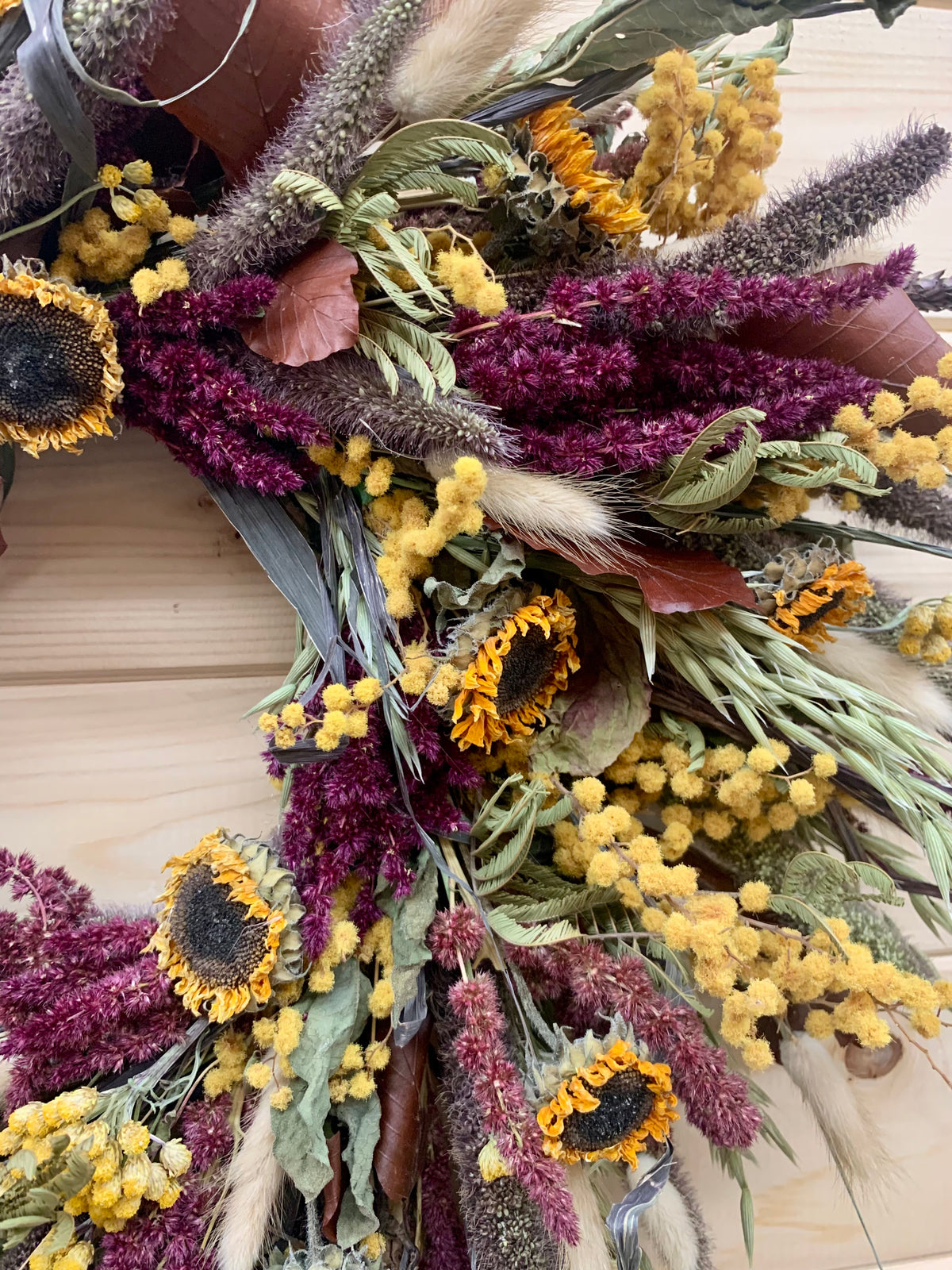 A rustic dried flower wreath featuring sunflowers, deep burgundy amaranthus, yellow mimosa, and natural red foxtail ornamental grasses on a light wood background.