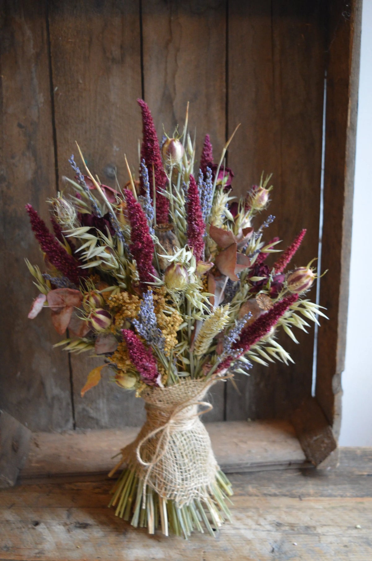 Bouquet of dried flowers in warm winter colours using burgundy amaranthus, love-in-a-mist seed heads, lavender and grasses tied with burlap against a wooden background