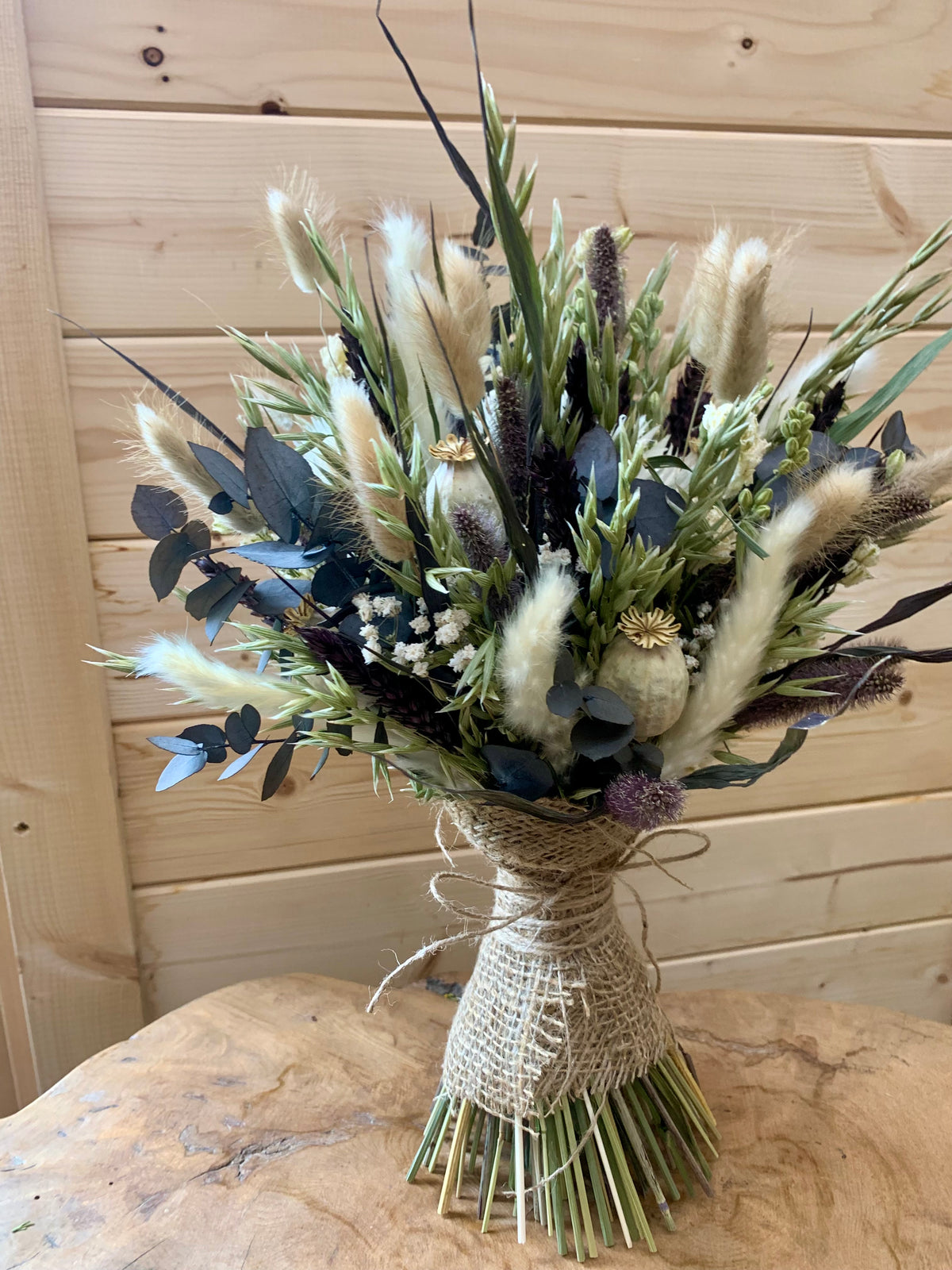 A dried floral bouquet featuring fluffy beige pampas grass, green and blue-green leaves, and small white flowers, wrapped in natural jute twine and tied with a bow, set on a rustic wooden surface against a light wood plank wall.