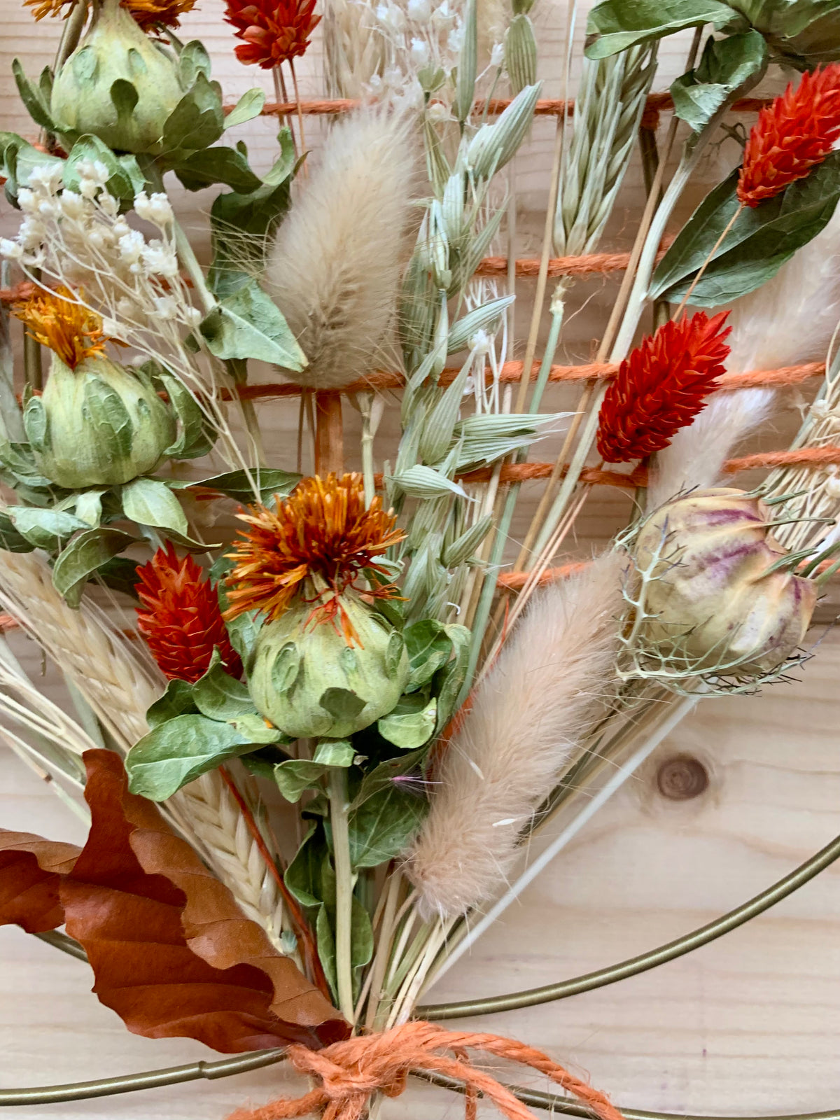 A close-up of a dried floral arrangement featuring orange-red flowers, green artichoke-like blooms, beige bunny tail grass, wheat stalks, and a brown leaf, tied with orange string against a light wood background.