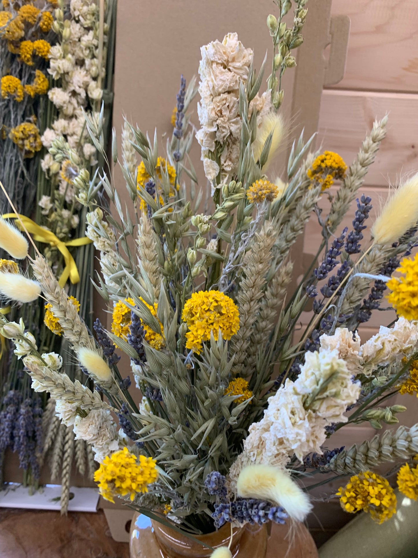A dried flower bouquet arrangement featuring yellow flowers, white and cream blossoms, lavender sprigs, and wheat stalks, photographed indoors against a light wood background.