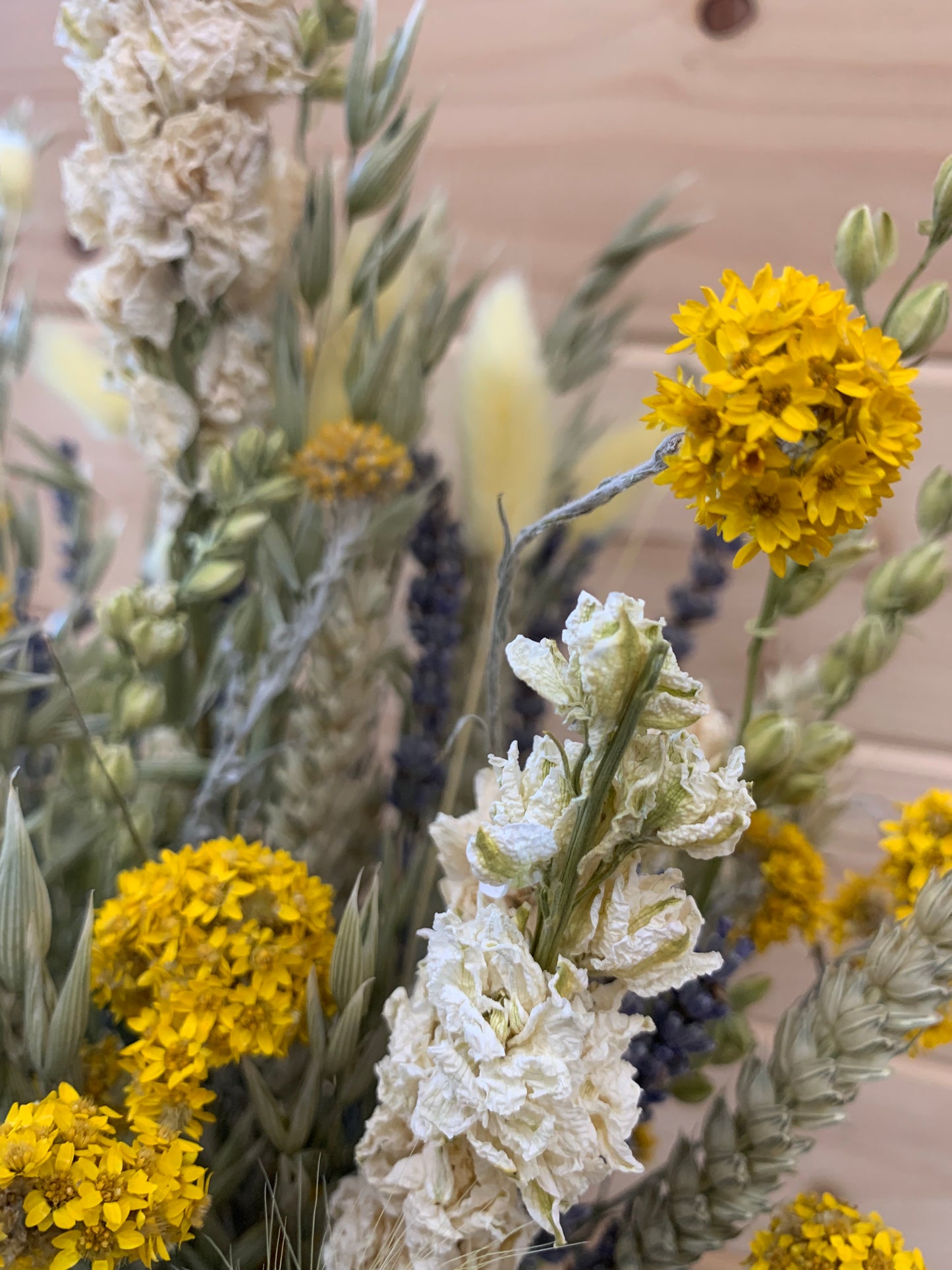 A close-up of a dried flower arrangement featuring bright yellow clusters, textured off-white ruffled blooms, and muted green stems against a soft pine background.