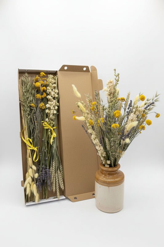 A rustic dried flower arrangement displayed in a stoneware jar next to the packing box including the same flowers  against a white background.
