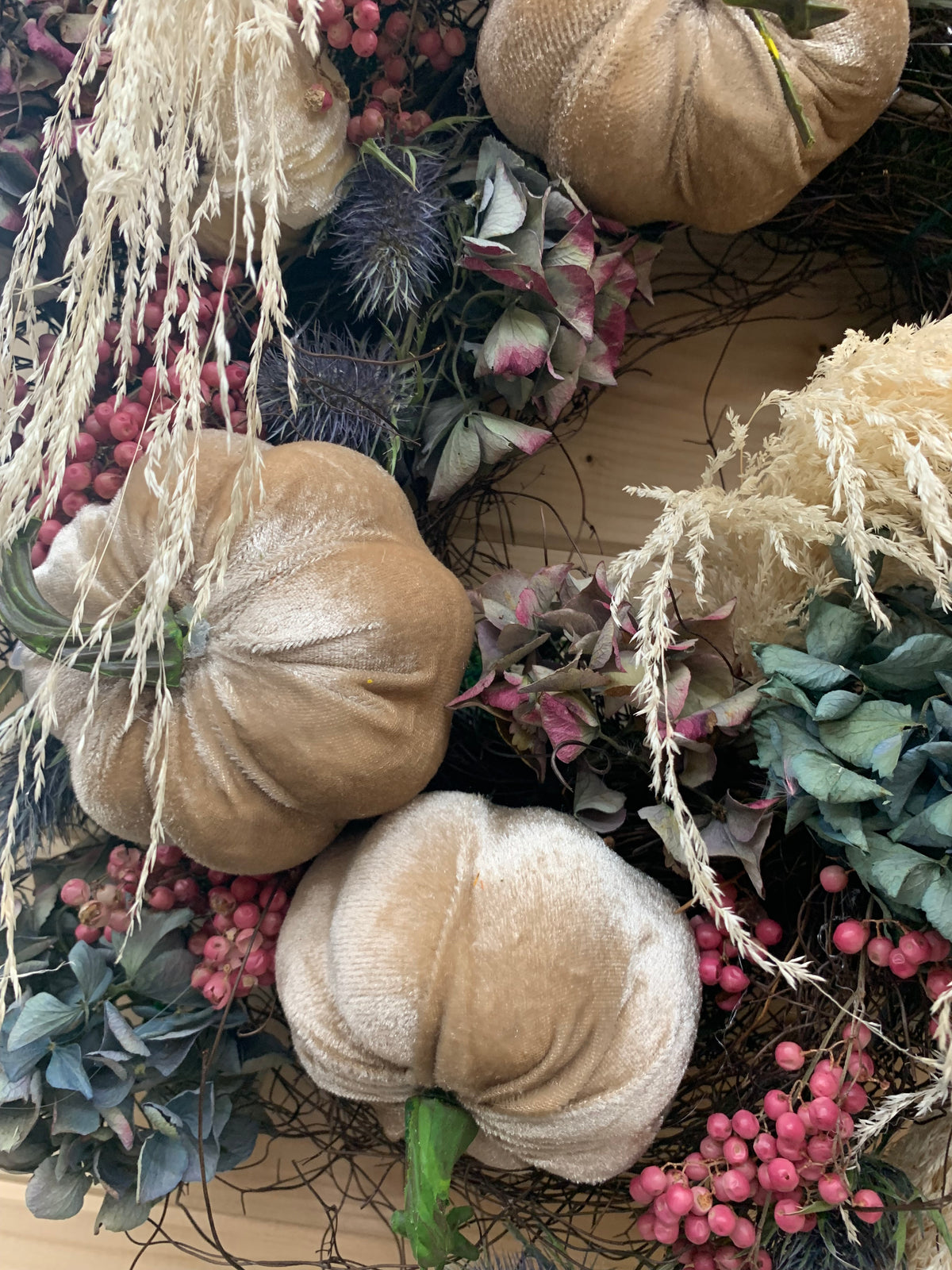 Autumn-themed decorative wreath featuring three faux velvet pumpkins in beige and cream shades, accented with pink pepper berries, dried hydrangea, and dried beige grasses.