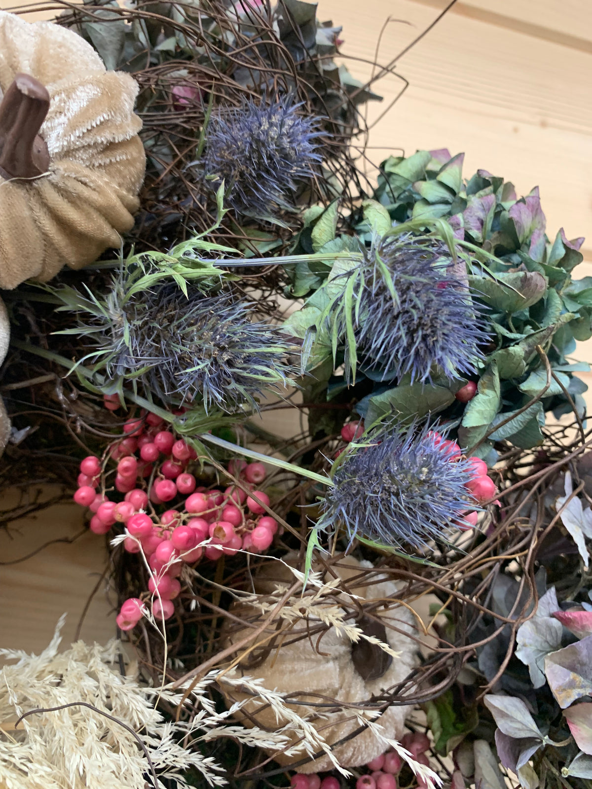A close-up of a rustic autumn wreath or floral arrangement featuring purple-blue thistle blooms, pink berries, green leaves, tan fabric pumpkin accents, and dried grasses on a light wood background.