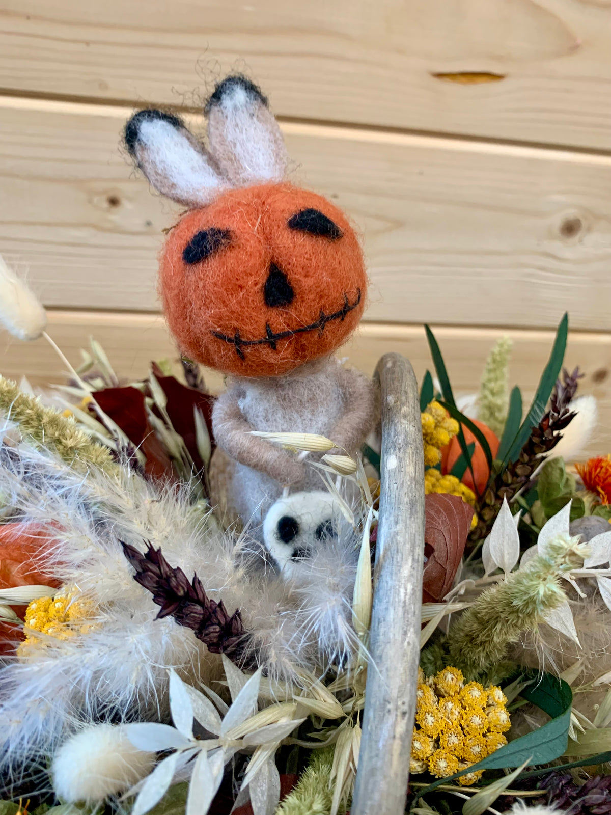 A handmade felted wool decorative arrangement featuring a small bunny figure with a carved orange jack-o'-lantern head, nestled among dried grasses, autumn leaves, and a driftwood branch against a light wood plank background.
